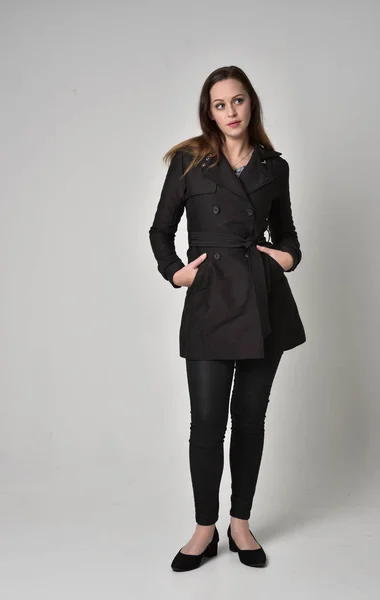 灰色のスタジオの背景に立ちポーズ長く黒いコートを着ているブルネットの少女の完全な長さの肖像画 — ストック写真