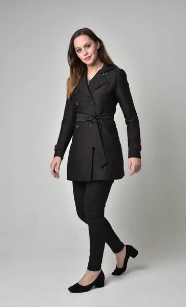 一个穿黑色长外套的黑发女孩的全长肖像 站在灰色的工作室背景摆姿势 — 图库照片