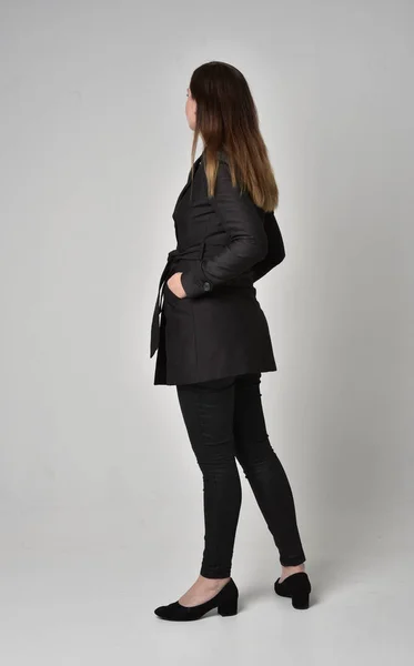 灰色のスタジオの背景にカメラに背中と立ちポーズ長く黒いコートを着ているブルネットの少女の完全な長さの肖像画 — ストック写真