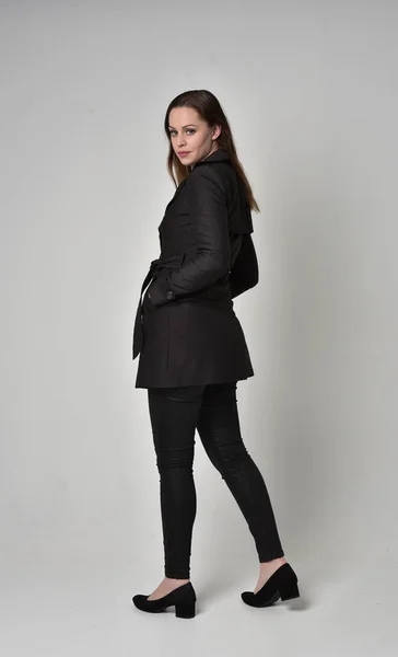 一个穿黑色长外套的黑发女孩的全长肖像 站在灰色工作室背景的镜头前摆姿势 — 图库照片