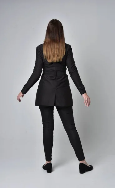 stock image full length portrait of a brunette girl wearing long black coat, standing pose with back to the camera on grey studio background.