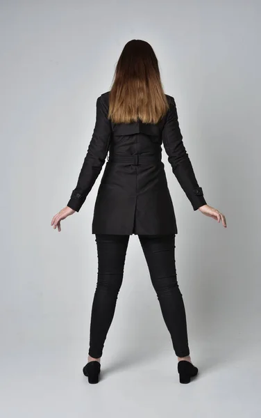 一个穿黑色长外套的黑发女孩的全长肖像 站在灰色工作室背景的镜头前摆姿势 — 图库照片