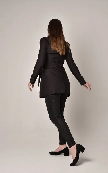 灰色のスタジオの背景にカメラに背中と立ちポーズ長く黒いコートを着ているブルネットの少女の完全な長さの肖像画 — ストック写真