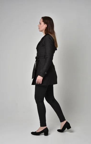 一个穿黑色长外套的黑发女孩的全长肖像 站在灰色工作室背景的侧面轮廓 — 图库照片