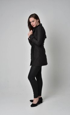 uzun, siyah ceket, gri studio arka plan üzerinde yan profil ayakta poz giyen bir esmer kız portresi tam uzunlukta.