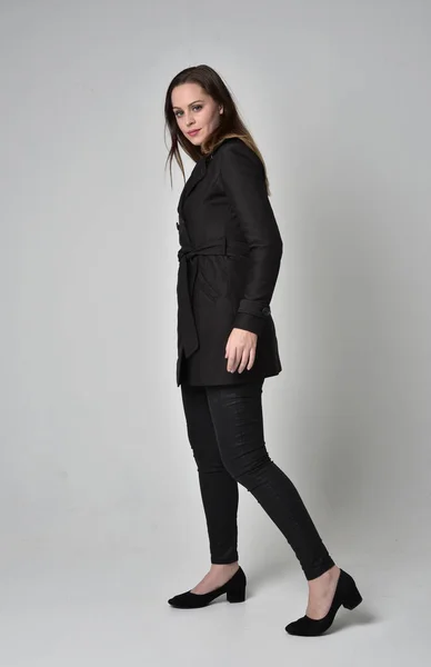 灰色のスタジオの背景に横顔の立ちポーズ長く黒いコートを着ているブルネットの少女の完全な長さの肖像画 — ストック写真