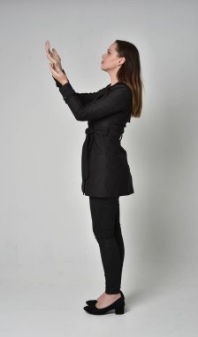 uzun, siyah ceket, gri studio arka plan üzerinde yan profil ayakta poz giyen bir esmer kız portresi tam uzunlukta.