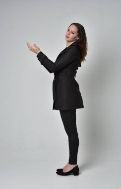 uzun, siyah ceket, gri studio arka plan üzerinde yan profil ayakta poz giyen bir esmer kız portresi tam uzunlukta.