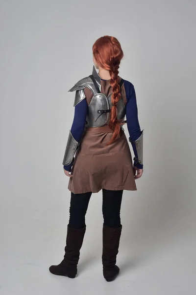 一个红头发的女孩穿着中世纪的战士服装和钢甲 站在站在灰色工作室背景的镜头前的姿势 — 图库照片