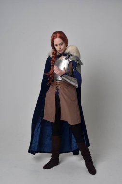 Ortaçağ savaşçı kostüm, mavi kadife pelerin ve çelik zırh, ayakta poz gri studio arka plan üzerinde kırmızı saçlı kız portresi tam uzunlukta.