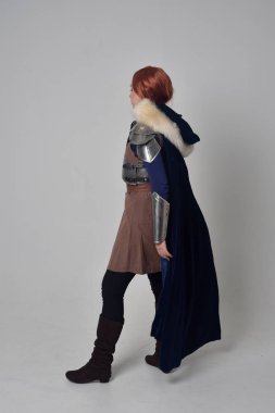 Ortaçağ savaşçı kostüm, mavi kadife pelerin ve çelik zırh, ayakta poz gri studio arka plan üzerinde kırmızı saçlı kız portresi tam uzunlukta.