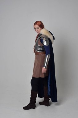 Ortaçağ savaşçı kostüm, mavi kadife pelerin ve çelik zırh, ayakta poz gri studio arka plan üzerinde kırmızı saçlı kız portresi tam uzunlukta.