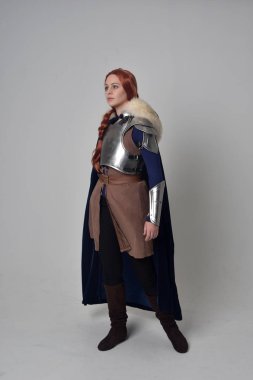 Ortaçağ savaşçı kostüm, mavi kadife pelerin ve çelik zırh, ayakta poz gri studio arka plan üzerinde kırmızı saçlı kız portresi tam uzunlukta.