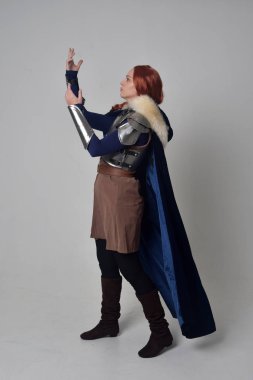 Ortaçağ savaşçı kostüm, mavi kadife pelerin ve çelik zırh, ayakta poz gri studio arka plan üzerinde kırmızı saçlı kız portresi tam uzunlukta.