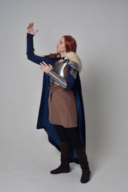 Ortaçağ savaşçı kostüm, mavi kadife pelerin ve çelik zırh, ayakta poz gri studio arka plan üzerinde kırmızı saçlı kız portresi tam uzunlukta.