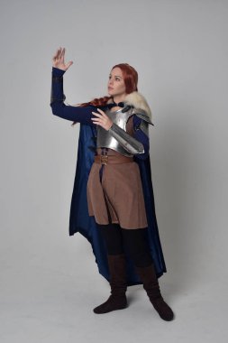 Ortaçağ savaşçı kostüm, mavi kadife pelerin ve çelik zırh, ayakta poz gri studio arka plan üzerinde kırmızı saçlı kız portresi tam uzunlukta.
