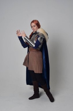 Ortaçağ savaşçı kostüm, mavi kadife pelerin ve çelik zırh, ayakta poz gri studio arka plan üzerinde kırmızı saçlı kız portresi tam uzunlukta.