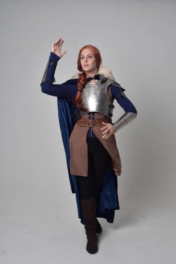 Ortaçağ savaşçı kostüm, mavi kadife pelerin ve çelik zırh, ayakta poz gri studio arka plan üzerinde kırmızı saçlı kız portresi tam uzunlukta.