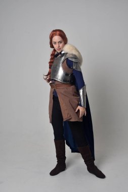 Ortaçağ savaşçı kostüm, mavi kadife pelerin ve çelik zırh, ayakta poz gri studio arka plan üzerinde kırmızı saçlı kız portresi tam uzunlukta.