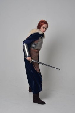 Ortaçağ savaşçı kostüm, mavi kadife pelerin ve çelik zırh, ayakta poz gri studio arka plan üzerinde kırmızı saçlı kız portresi tam uzunlukta.