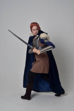 Ortaçağ savaşçı kostüm, mavi kadife pelerin ve çelik zırh, ayakta poz gri studio arka plan üzerinde kırmızı saçlı kız portresi tam uzunlukta.