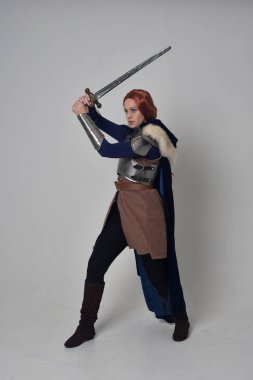 Ortaçağ savaşçı kostüm, mavi kadife pelerin ve çelik zırh, ayakta poz gri studio arka plan üzerinde kırmızı saçlı kız portresi tam uzunlukta.