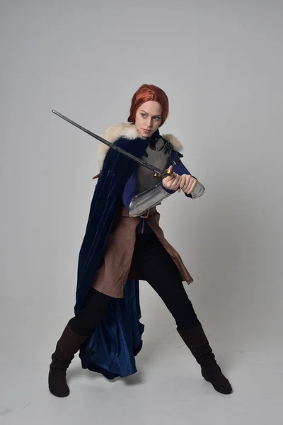 一个红头发的女孩穿着中世纪的战士服装 蓝色天鹅绒斗篷和钢甲 站在灰色的工作室背景的姿势的全长肖像 — 图库照片
