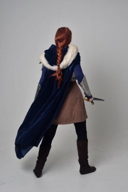 Ortaçağ savaşçı kostüm, mavi kadife pelerin ve çelik zırh, ayakta poz gri studio arka plan üzerinde kırmızı saçlı kız portresi tam uzunlukta.