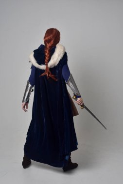 Ortaçağ savaşçı kostüm, mavi kadife pelerin ve çelik zırh, ayakta poz gri studio arka plan üzerinde kırmızı saçlı kız portresi tam uzunlukta.