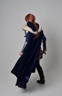 Ortaçağ savaşçı kostüm, mavi kadife pelerin ve çelik zırh, ayakta poz gri studio arka plan üzerinde kırmızı saçlı kız portresi tam uzunlukta.