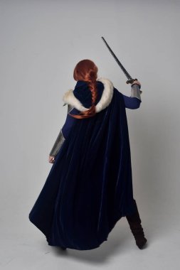 Ortaçağ savaşçı kostüm, mavi kadife pelerin ve çelik zırh, ayakta poz gri studio arka plan üzerinde kırmızı saçlı kız portresi tam uzunlukta.