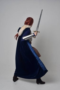 Ortaçağ savaşçı kostüm, mavi kadife pelerin ve çelik zırh, ayakta poz gri studio arka plan üzerinde kırmızı saçlı kız portresi tam uzunlukta.