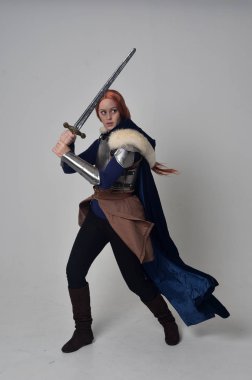 Ortaçağ savaşçı kostüm, mavi kadife pelerin ve çelik zırh, ayakta poz gri studio arka plan üzerinde kırmızı saçlı kız portresi tam uzunlukta.