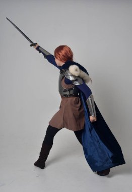 Ortaçağ savaşçı kostüm, mavi kadife pelerin ve çelik zırh, ayakta poz gri studio arka plan üzerinde kırmızı saçlı kız portresi tam uzunlukta.