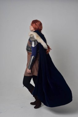 Ortaçağ savaşçı kostüm, mavi kadife pelerin ve çelik zırh, ayakta poz gri studio arka plan üzerinde kırmızı saçlı kız portresi tam uzunlukta.