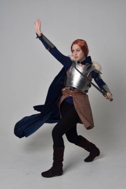 Ortaçağ savaşçı kostüm, mavi kadife pelerin ve çelik zırh, ayakta poz gri studio arka plan üzerinde kırmızı saçlı kız portresi tam uzunlukta.