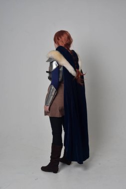 Ortaçağ savaşçı kostüm, mavi kadife pelerin ve çelik zırh, ayakta poz gri studio arka plan üzerinde kırmızı saçlı kız portresi tam uzunlukta.