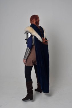 Ortaçağ savaşçı kostüm, mavi kadife pelerin ve çelik zırh, ayakta poz gri studio arka plan üzerinde kırmızı saçlı kız portresi tam uzunlukta.