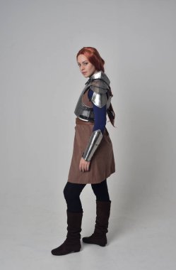 Ortaçağ savaşçı kostüm ve çelik zırh, ayakta poz gri studio arka plan üzerinde kırmızı saçlı kız portresi tam uzunlukta.