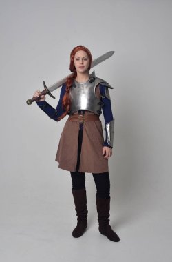 tam uzunlukta bir kılıç tutan Ortaçağ savaşçı kostüm ve çelik zırh giyen bir kırmızı saçlı kız portresi. gri studio arka plan üzerinde ayakta poz.