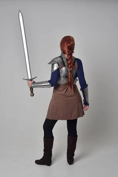一个红头发的女孩穿着中世纪的战士服装和钢甲 拿着一把剑的全长肖像 站在灰色工作室背景的姿势 — 图库照片