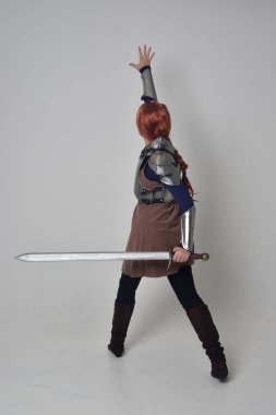 tam uzunlukta bir kılıç tutan Ortaçağ savaşçı kostüm ve çelik zırh giyen bir kırmızı saçlı kız portresi. gri studio arka plan üzerinde ayakta poz.