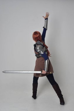 tam uzunlukta bir kılıç tutan Ortaçağ savaşçı kostüm ve çelik zırh giyen bir kırmızı saçlı kız portresi. gri studio arka plan üzerinde ayakta poz.
