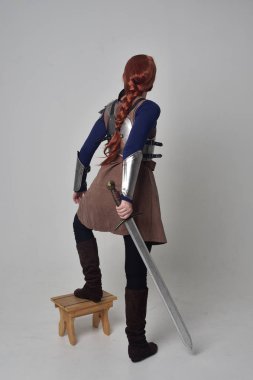 tam uzunlukta bir kılıç tutan Ortaçağ savaşçı kostüm ve çelik zırh giyen bir kırmızı saçlı kız portresi. gri studio arka plan üzerinde ayakta poz.