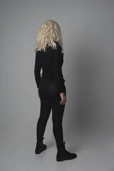 金髪少女をモダンな黒いジャケットとズボンを身に着けている灰色のスタジオの背景に立ちポーズの完全な長さの肖像画 — ストック写真
