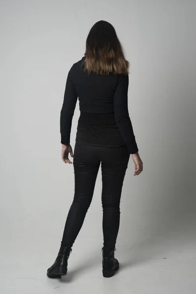 ブルネットの少女モダンな黒いジャケットとズボンを身に着けている灰色のスタジオの背景にカメラに背中と立ちポーズの完全な長さの肖像画 — ストック写真