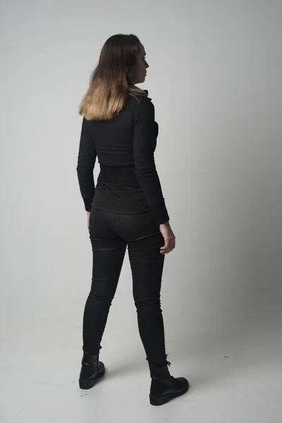 ブルネットの少女モダンな黒いジャケットとズボンを身に着けている灰色のスタジオの背景にカメラに背中と立ちポーズの完全な長さの肖像画 — ストック写真