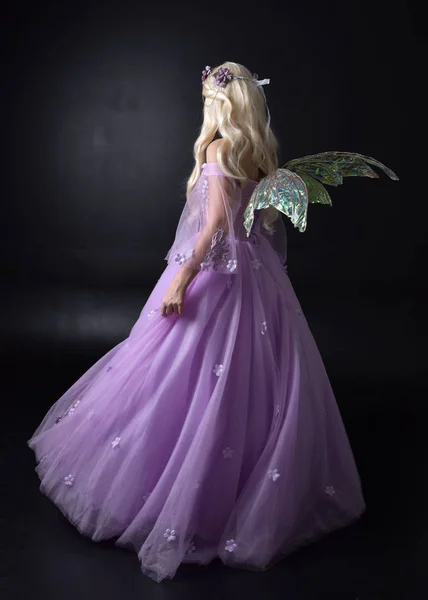 full length portrait of a blonde girl wearing a fantasy fairy inspired costume,  long purple ball gown with fairy wings,   standing pose  with back to the camera on a dark studio background.