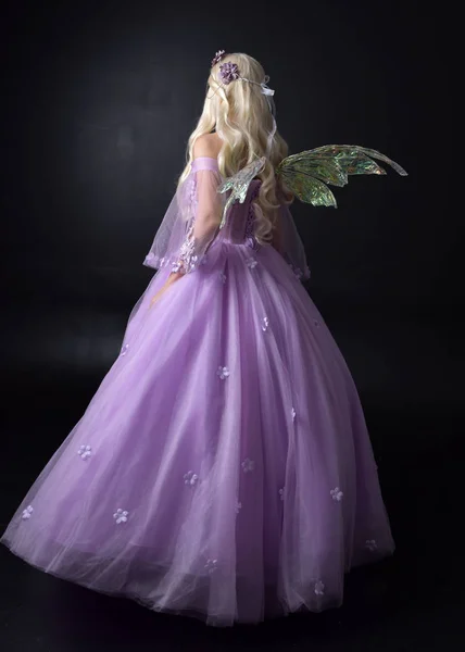full length portrait of a blonde girl wearing a fantasy fairy inspired costume,  long purple ball gown with fairy wings,   standing pose  with back to the camera on a dark studio background.