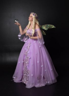 full length portrait of a blonde girl wearing a fantasy fairy inspired costume,  long purple ball gown with fairy wings,   sitting pose  on a dark studio background. clipart